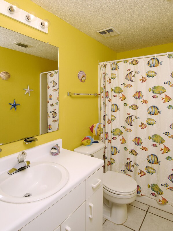 A bathroom with a yellow wall, white fixtures, a mirror, and a shower curtain with fish patterns. - Tidewater By Wyndham Vacation Rentals (Orange Beach)