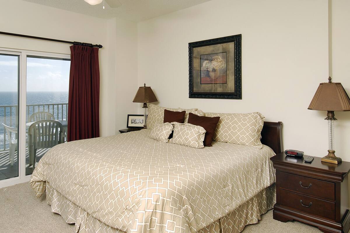 A bedroom with a bed, a nightstand, a lamp, a framed picture, and a view of the ocean.