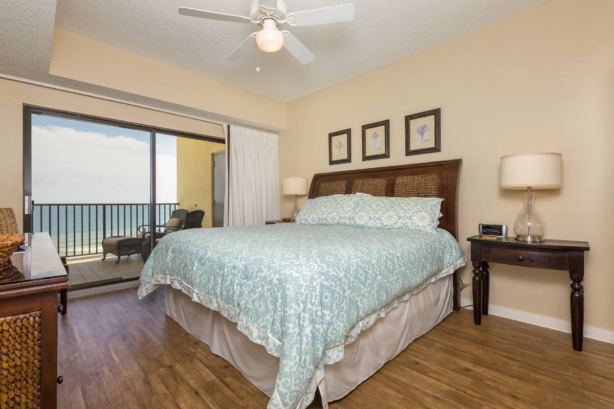 Habitación de hotel con una cama grande, un ventilador de techo, una mesita de noche y vista al océano a través de una puerta corrediza.