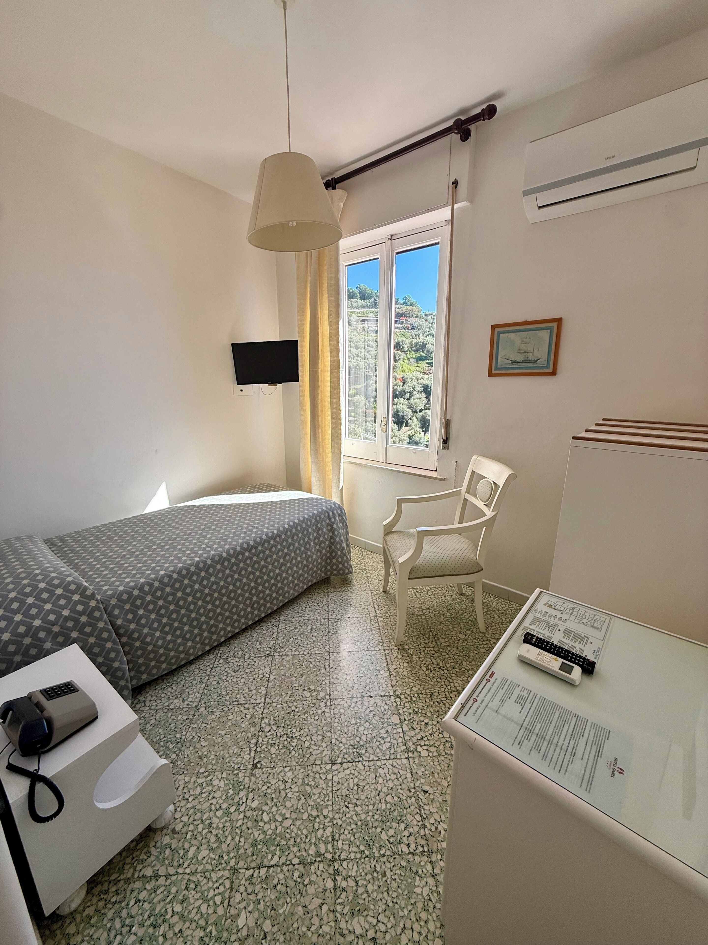 A room with a bed, a desk, a chair, a TV, a window with a view, and an air conditioner.