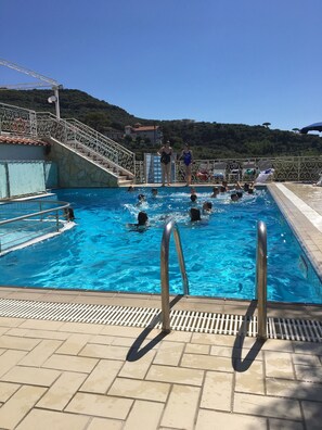 Una piscina con gente nadando, una escalera de metal y un área de terrazas con baldosas.