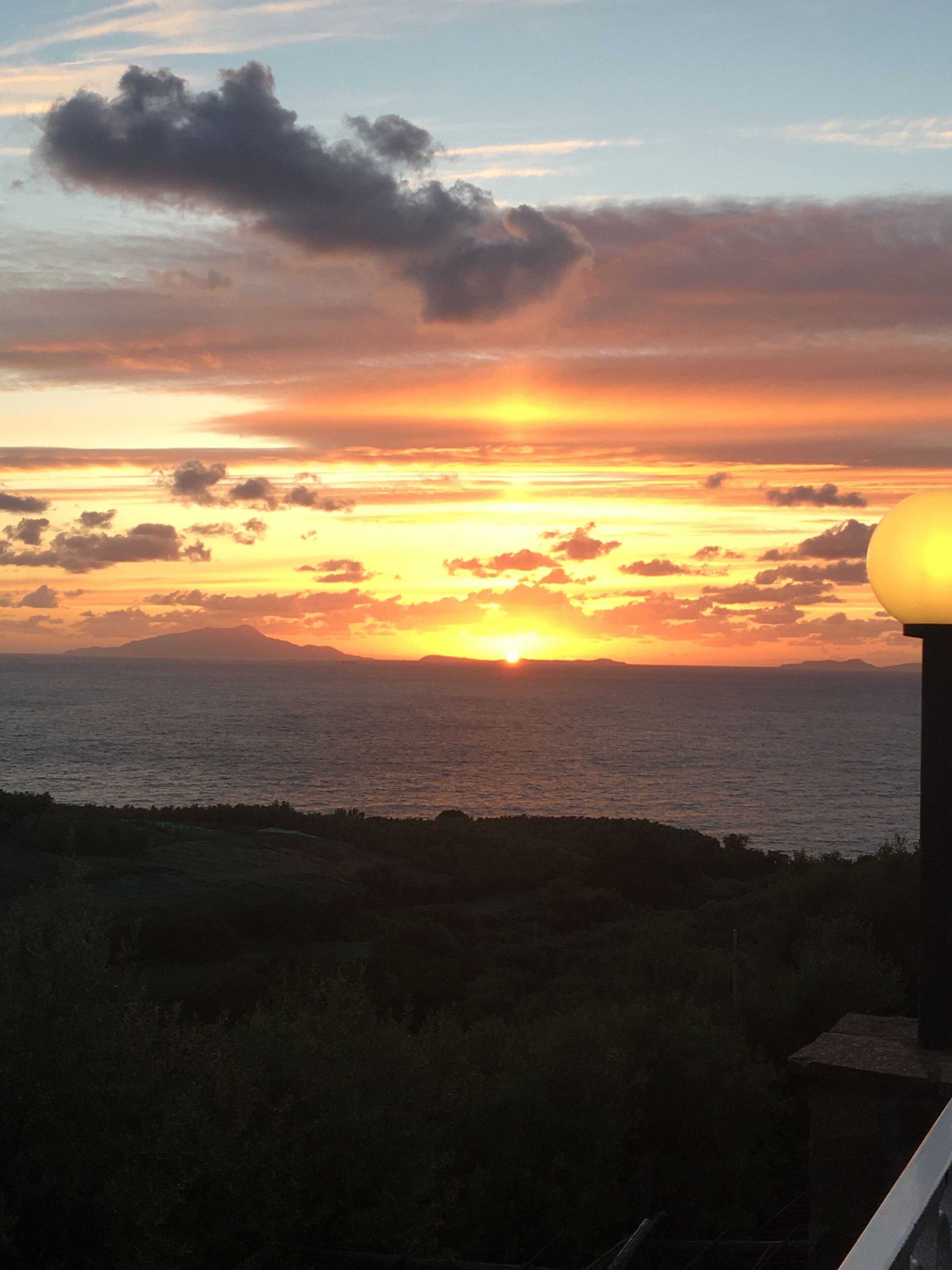 Um pôr do sol sobre o oceano, com o perfil da costa e um poste de luz ao primeiro plano.