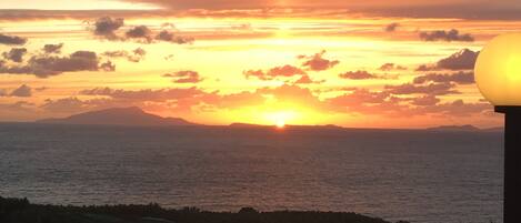 Um pôr do sol sobre o oceano, com o perfil da costa e um poste de luz ao primeiro plano.