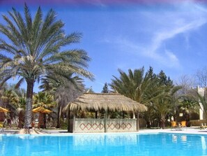 A swimming pool surrounded by palm trees and a thatched-roof structure. - Hotel Sahara Douz (Douz Nord)
