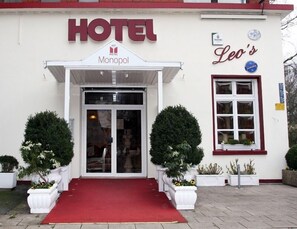 Entrada de un hotel con alfombra roja, dos plantas en macetas y un cartel que dice 'HOTEL Monopol Leo's'.