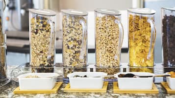 A breakfast bar with five glass containers filled with different types of cereal and two white dishes with spoons.