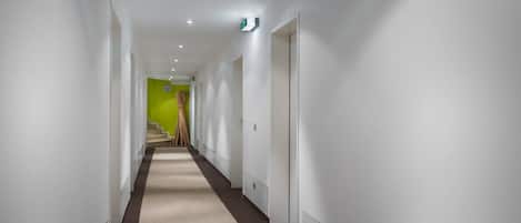 Un pasillo moderno de hotel con paredes blancas, alfombra color beige y una pared de acento verde al final.