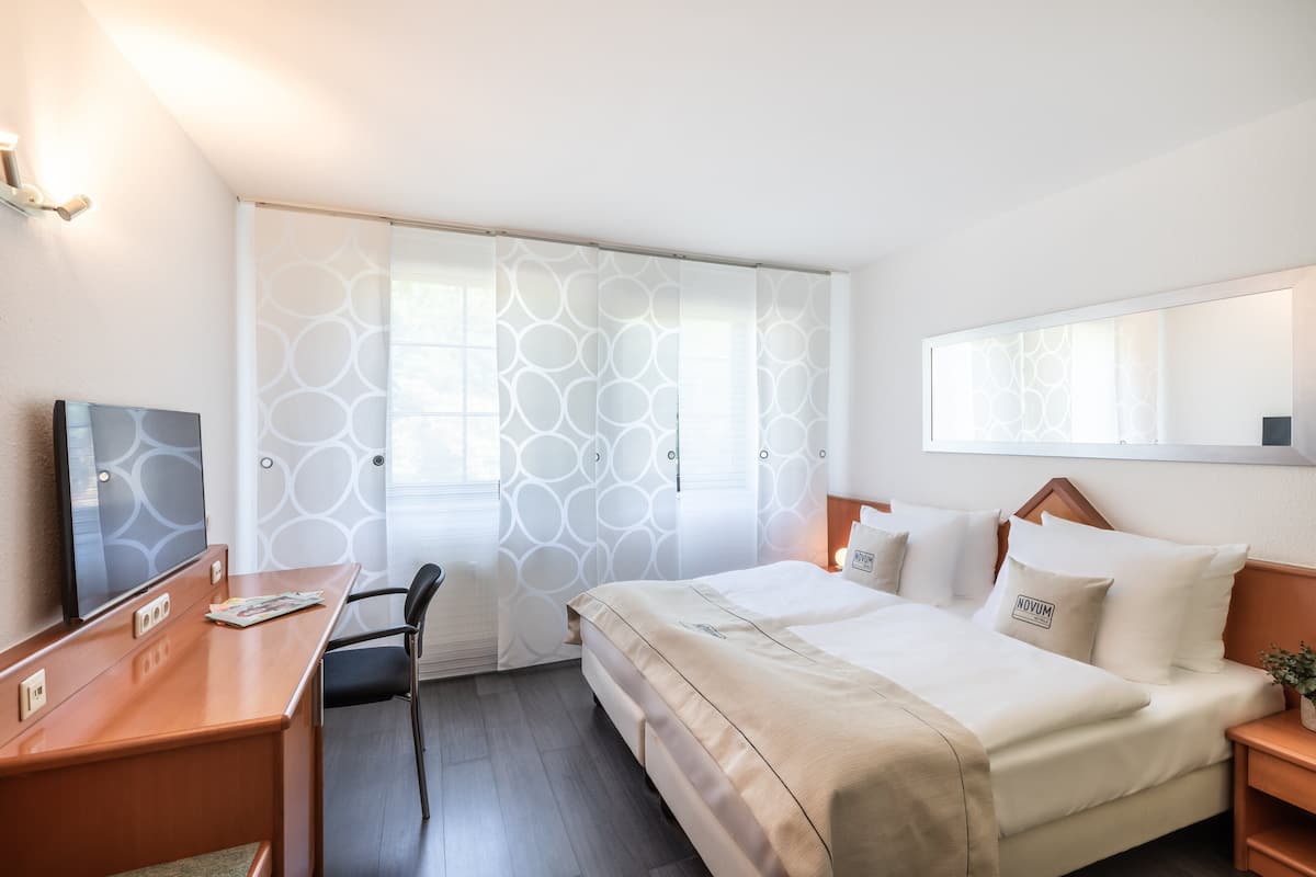 Une chambre d’hôtel avec un lit, un bureau, une chaise, une télévision et une fenêtre avec des rideaux à motifs.