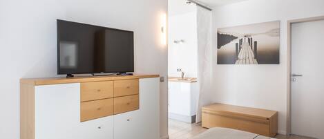 Un dormitorio moderno con televisión sobre un mueble de madera, cama con manta de marca, mesita de noche pequeña de madera y un cuadro de un muelle en la pared.