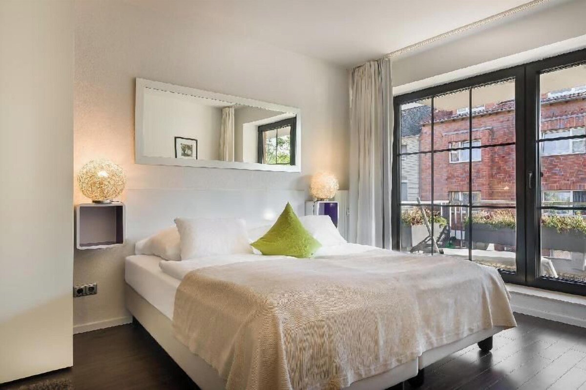 A hotel room with a bed, a large window, a mirror, and a wall-mounted shelf.