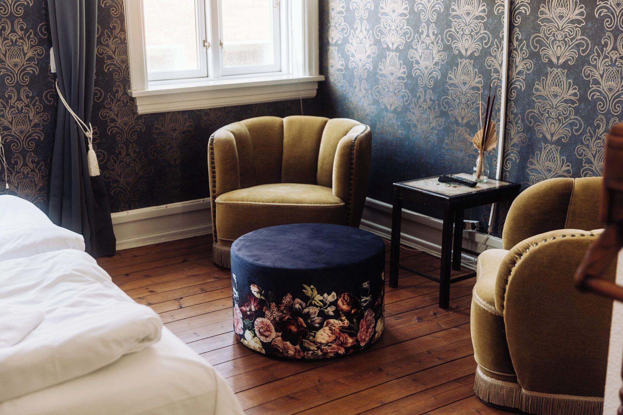 Ein Zimmer mit einem Bett, einem runden Ottomanen mit Blumenmuster, einem Sessel und einem kleinen Tisch mit Fernbedienung und Dekoration.