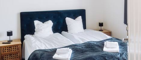 Una cama bien hecha con sábanas blancas y un cabecero oscuro, una mesita de noche de madera con una lámpara, y una alfombra azul con motivos de estrellas.