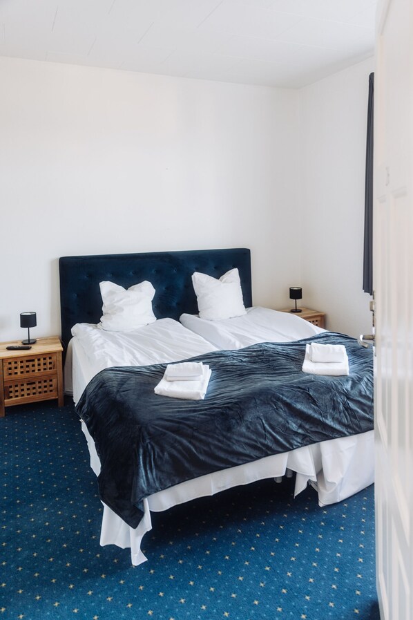 A neatly made bed with white linens and a dark headboard, a wooden nightstand with a lamp, and a blue carpet with star patterns.