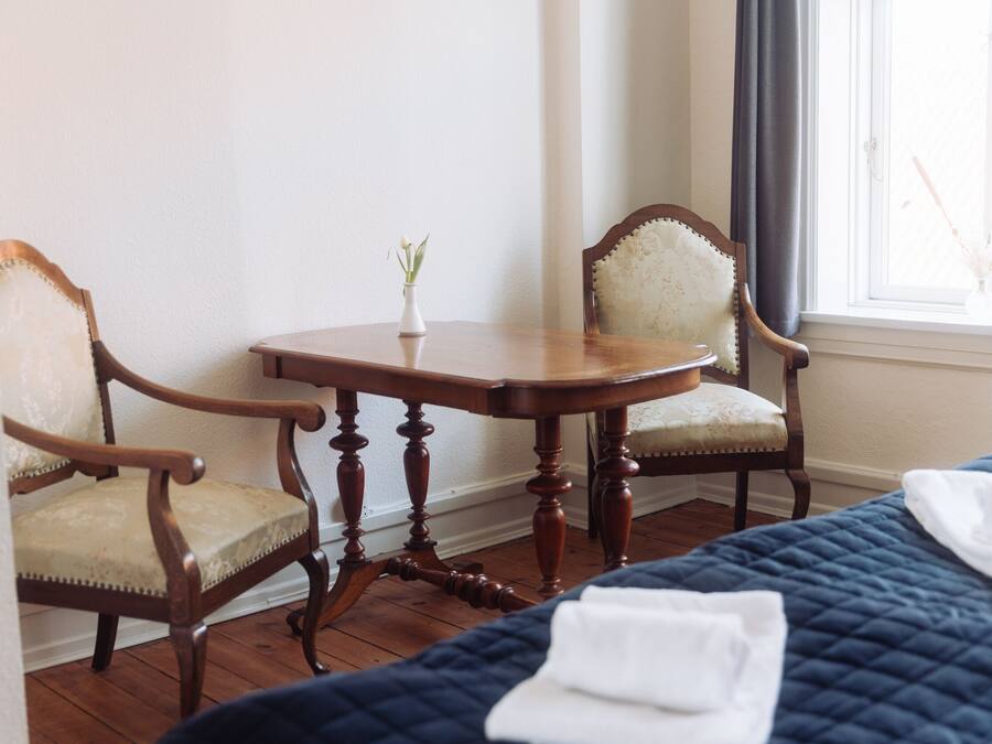 Una stanza con due sedie di legno, un tavolino con un vaso e un letto con copriletto trapuntato.