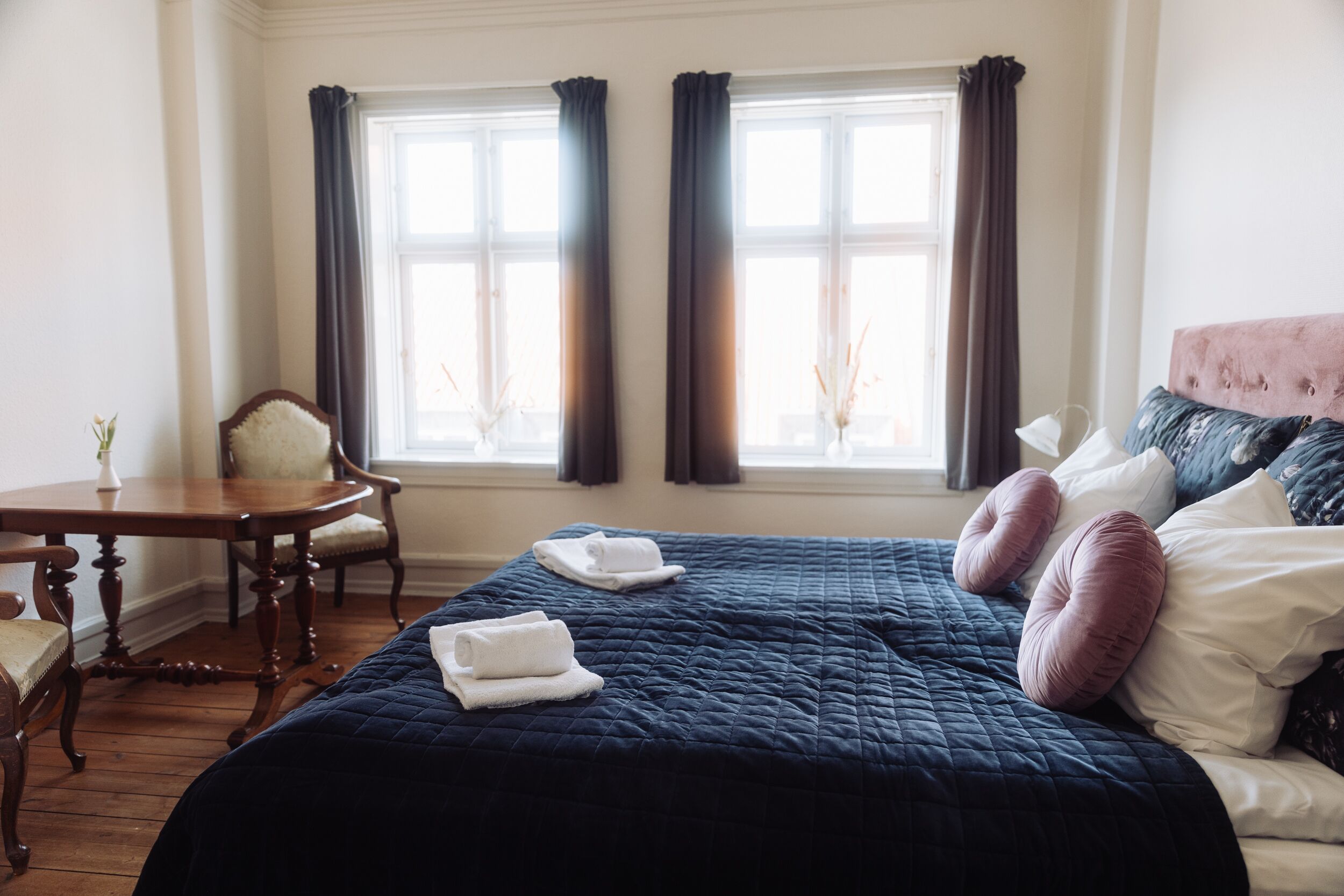 Un dormitorio con una cama grande, dos ventanas con cortinas, una silla de madera y una mesita con un florero.