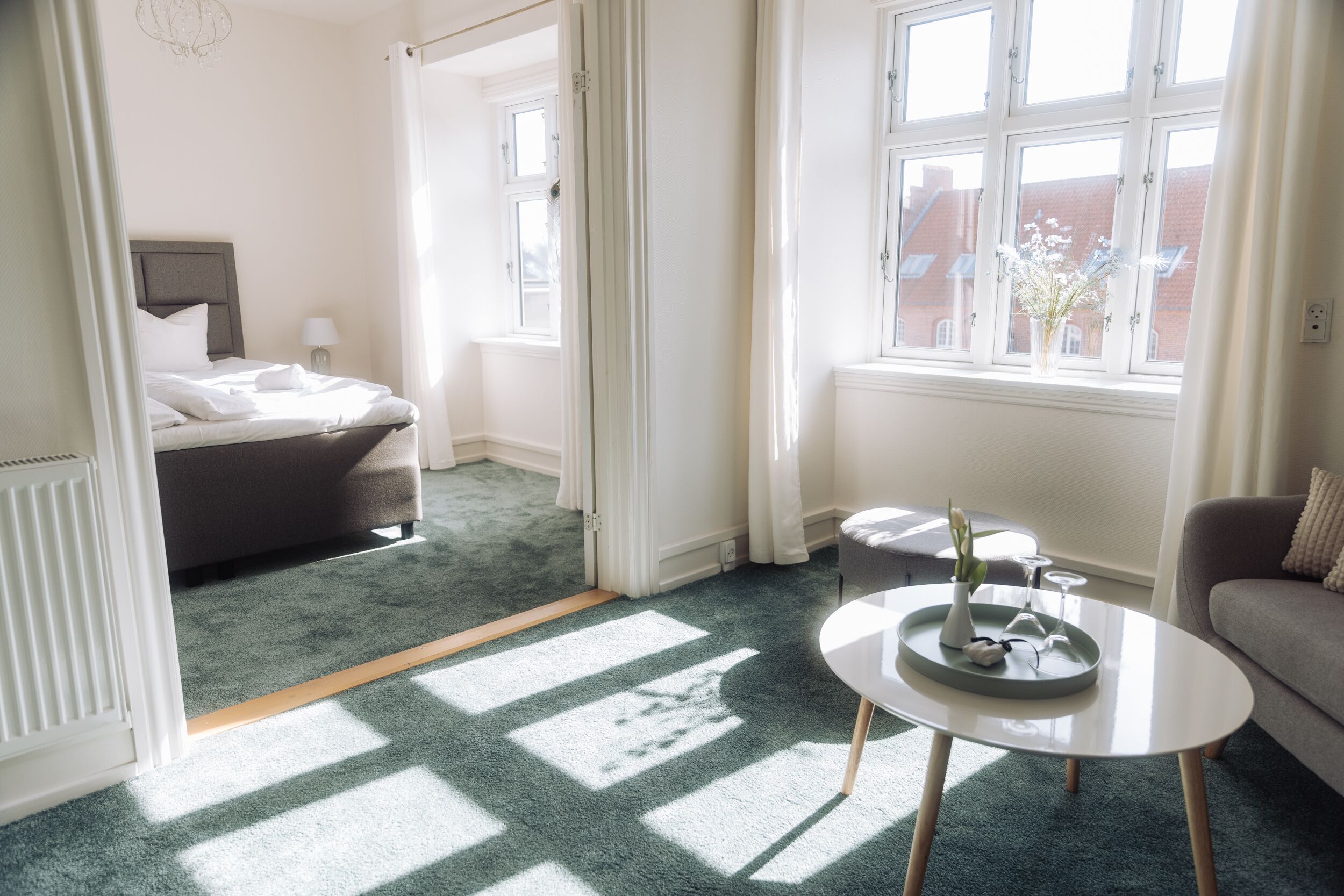 Quarto de hotel moderno com uma cama, uma mesinha com um vaso e copos, um sofá e janelas amplas com luz natural.