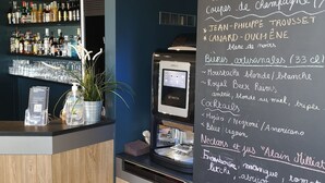 Un área de bar con un menú en pizarra que enumera diversas bebidas y aperitivos, una máquina de café KREA y una estantería con bebidas embotelladas.