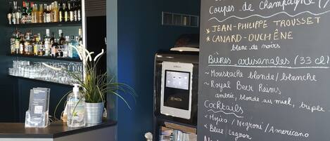 A bar area with a chalkboard menu listing various drinks and snacks, a KREA coffee machine, and a shelf of bottled beverages.
