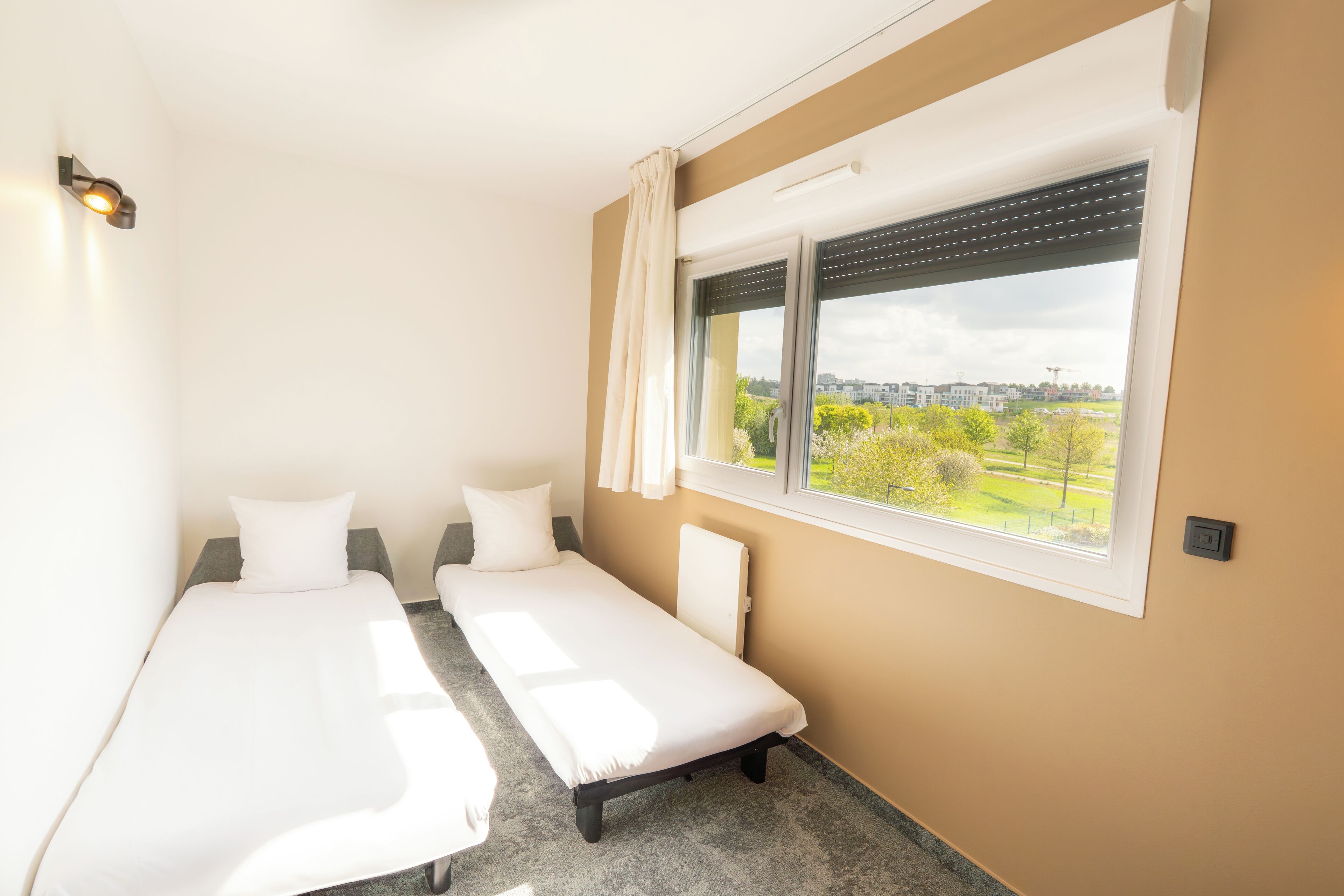 Een kleine hotelkamer met één ruimte, twee eenpersoonsbedden, een raam met uitzicht op een stadsbeeld en een wandlamp.