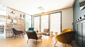 Un interior moderno con piso de madera, varios sillones, una estantería y ventanas amplias con persianas.