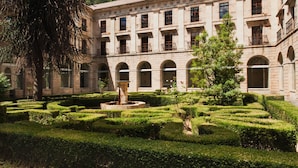 Un patio con una fuente, setos cuidados y un edificio con ventanas arqueadas y balcones.