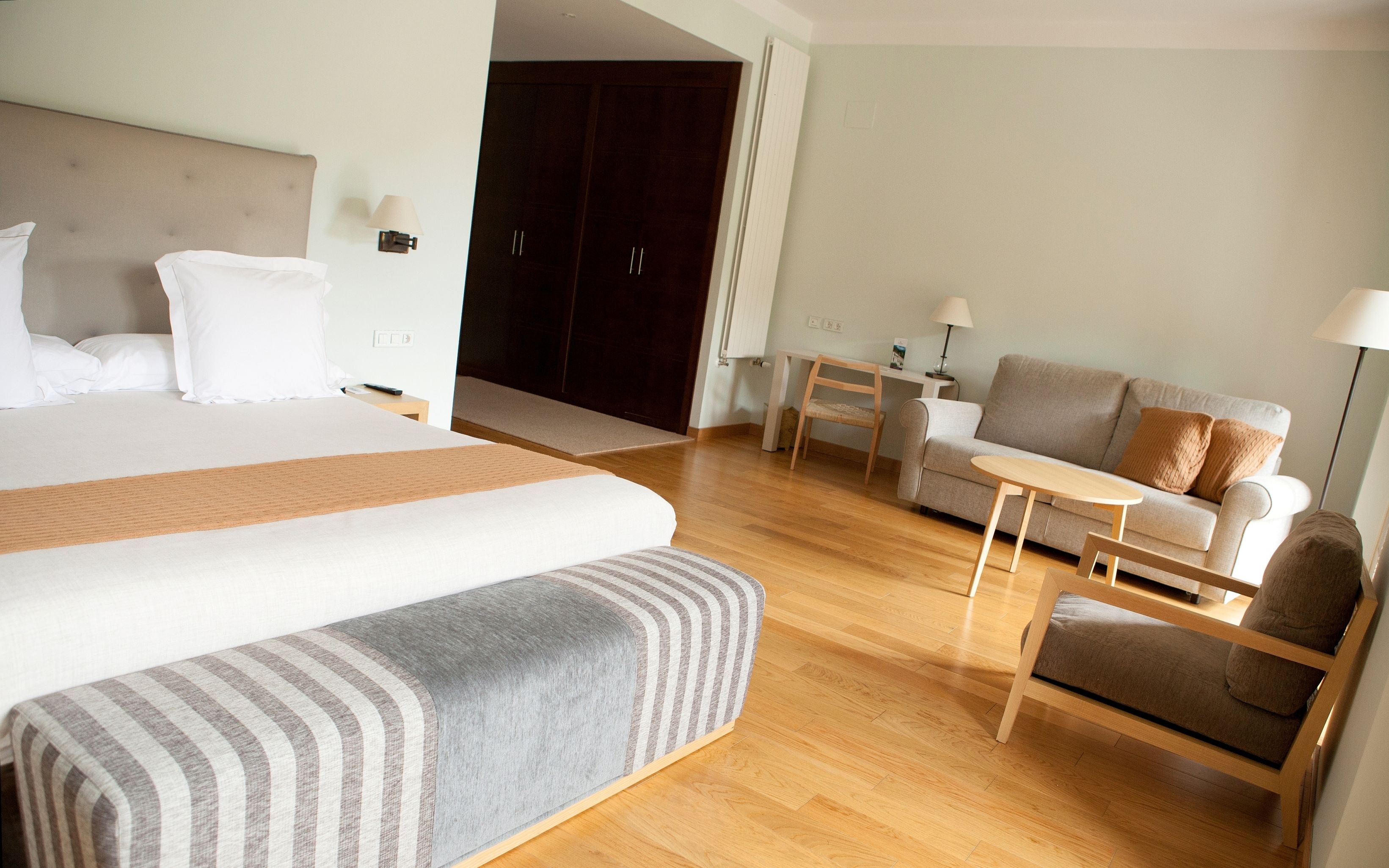 Habitación de hotel con una cama grande, un sofá, una mesita y una silla. Se ve una puerta que conduce a otra habitación y una ventana con cortinas.