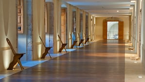 Un largo pasillo con bancos de madera y pilares de piedra, que conduce a una habitación luminosa con un amplio ventanal.
