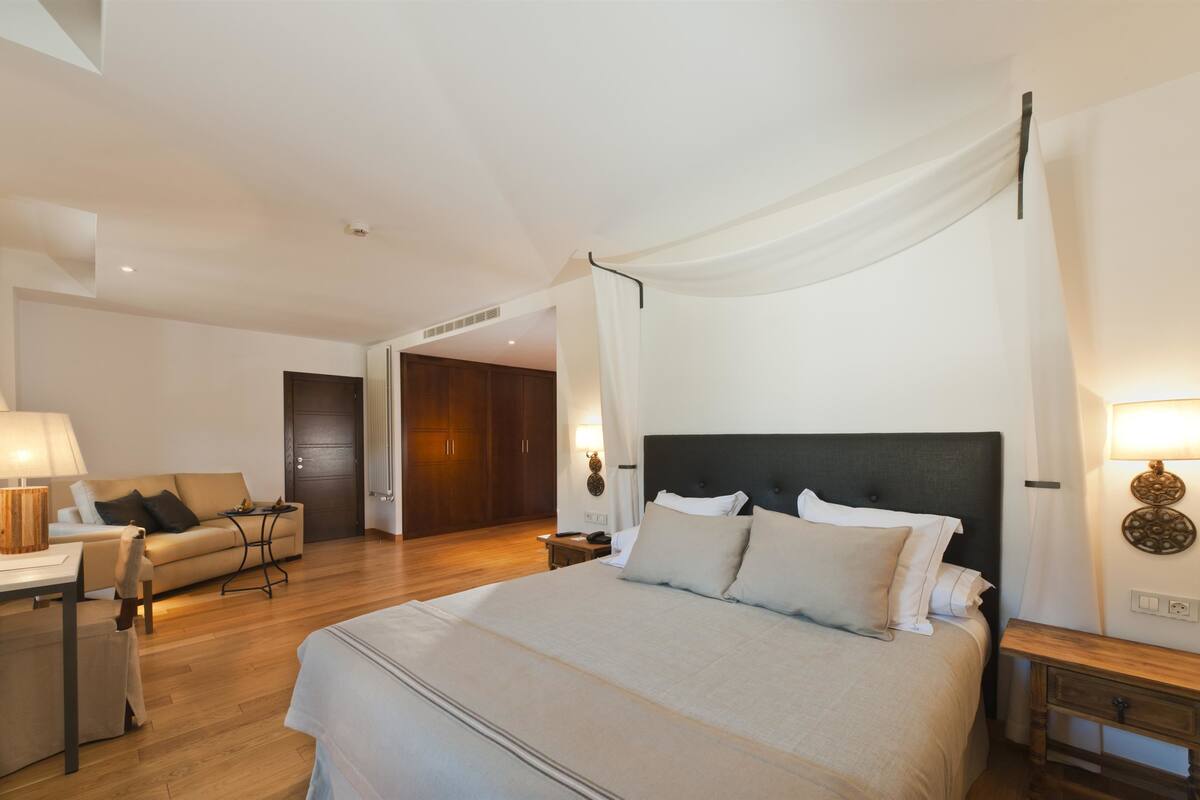 Une chambre à coucher avec un grand lit, une table de chevet, une lampe, un plancher en bois, une porte et un luminaire fixé au mur.