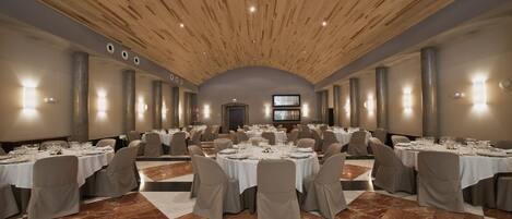 Un amplio salón de banquetes con varias mesas redondas dispuestas para comer, con un alto techo de madera y suelo de mármol.