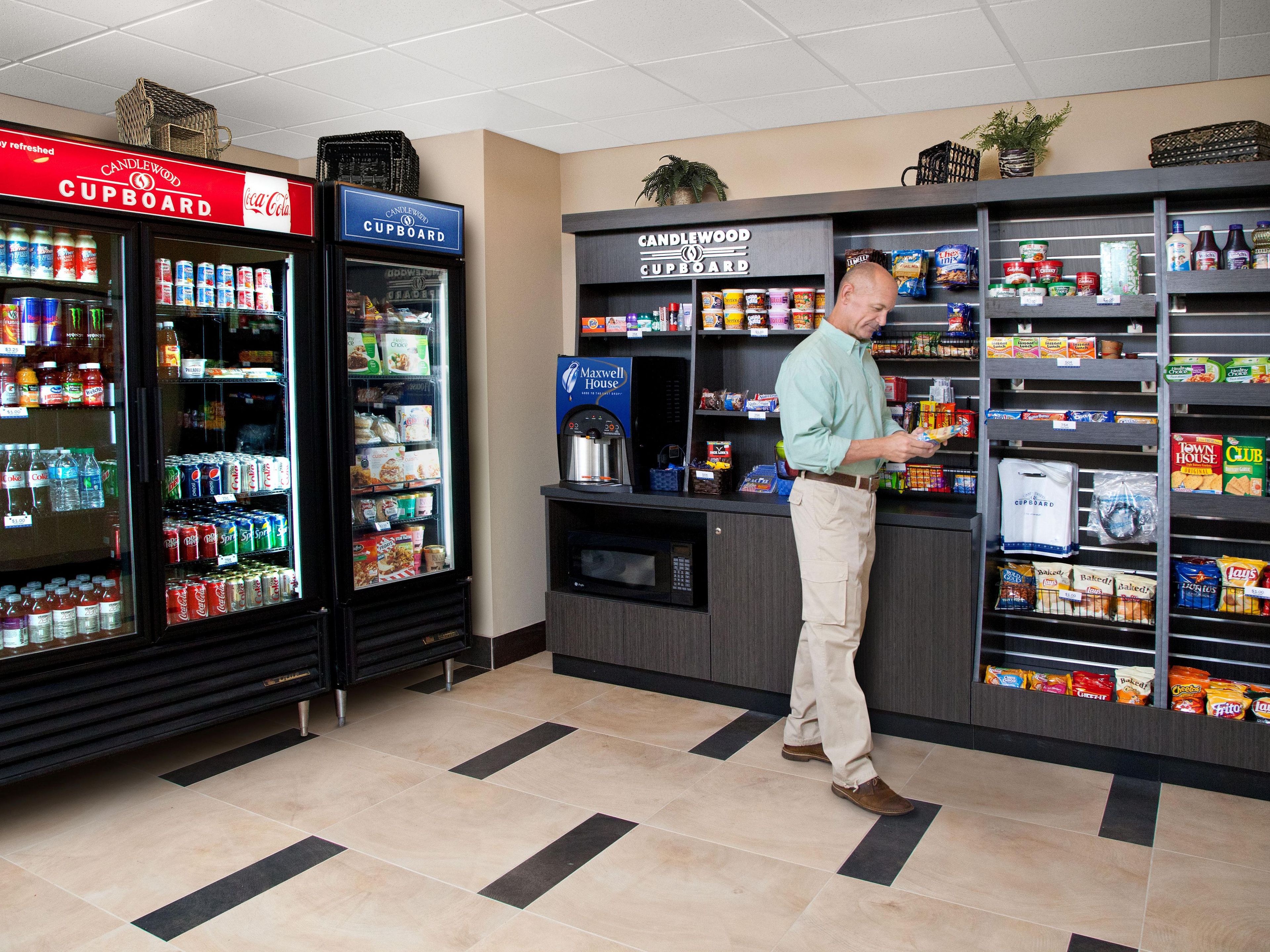 A man in a kitchenette area with a refrigerator stocked with beverages, a coffee machine, and shelves with snacks and condiments.