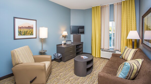 Quarto de hotel com uma poltrona bege, um sofá marrom com almofadas listradas, uma mesinha redonda, uma TV de tela plana em um suporte escuro, uma escrivaninha com abajur, um quadro na parede e cortinas amarelas.