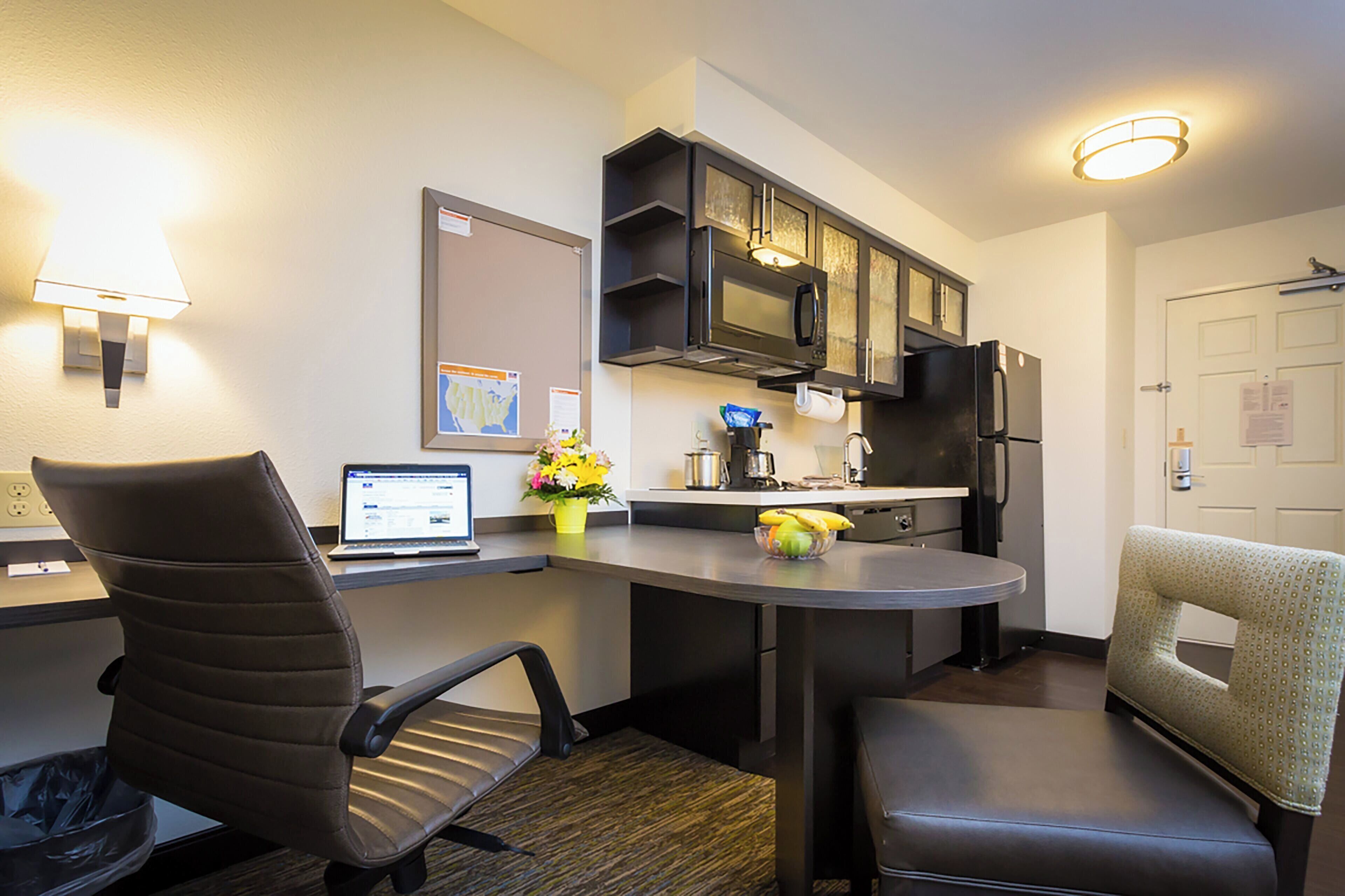 A compact kitchen with a microwave, refrigerator, and a laptop on the counter. There are two chairs and a small table.