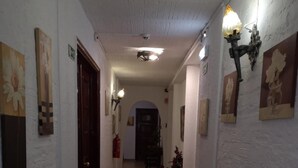 Un corridoio con pareti bianche, pavimento piastrellato e lampade a parete. Si vedono quadri incorniciati e un estintore.
