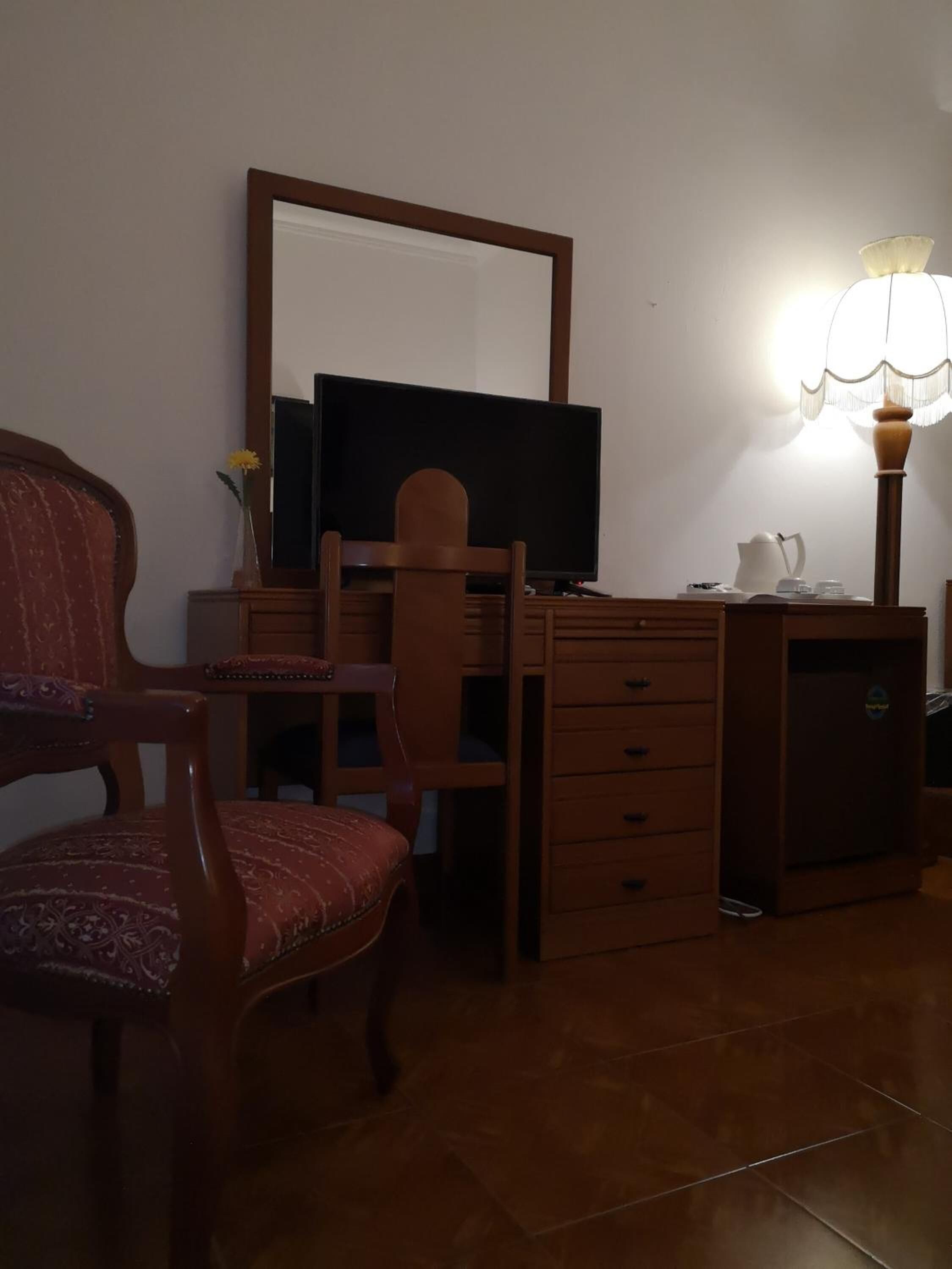 Quarto com escrivaninha de madeira, cadeira, televisão, luminária e um pequeno frigorífico.