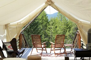 A tent with two wooden chairs and a table, set against a backdrop of trees and mountains.