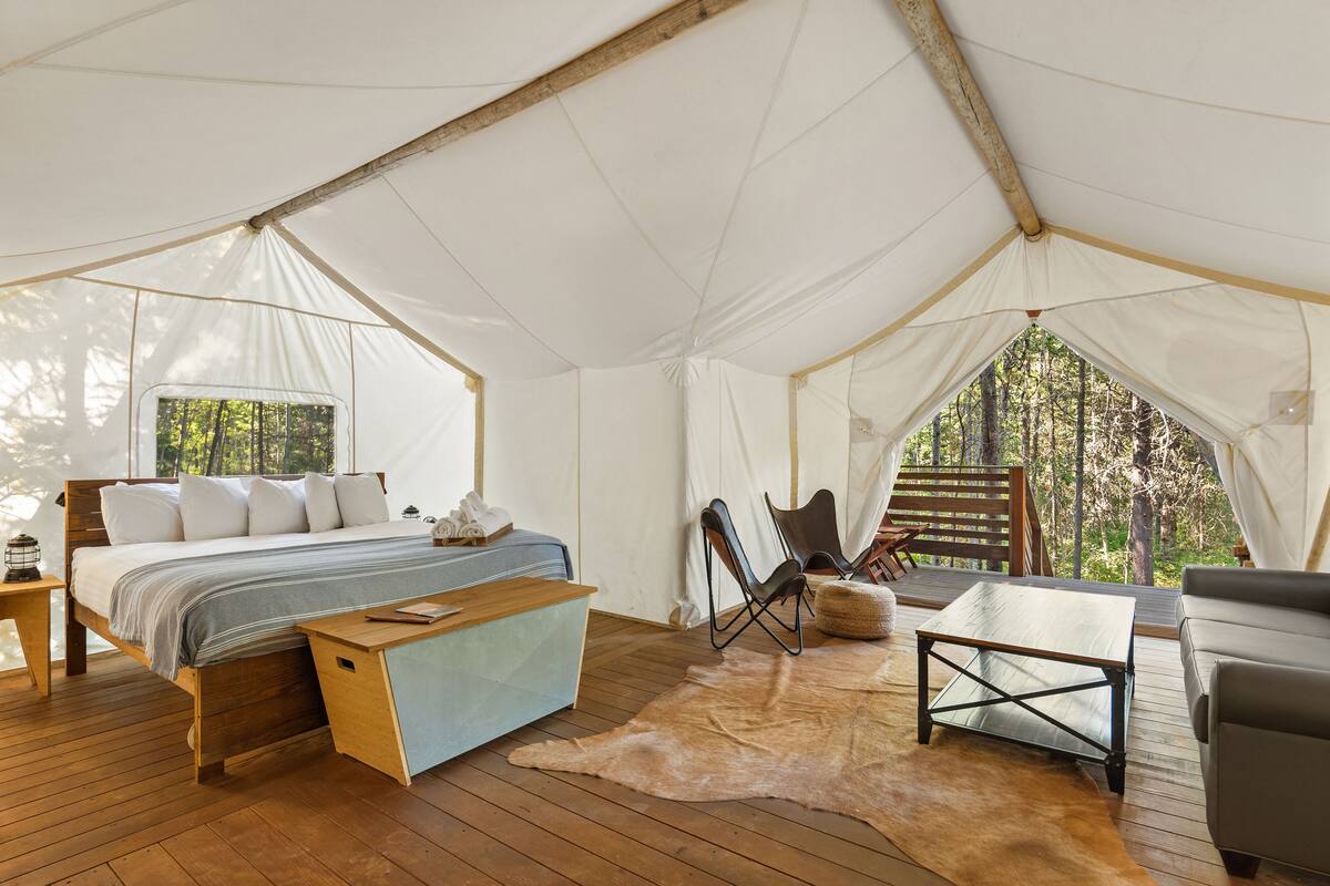 Alojamiento en tienda de campaña con cama, escritorio, silla y vista al bosque.