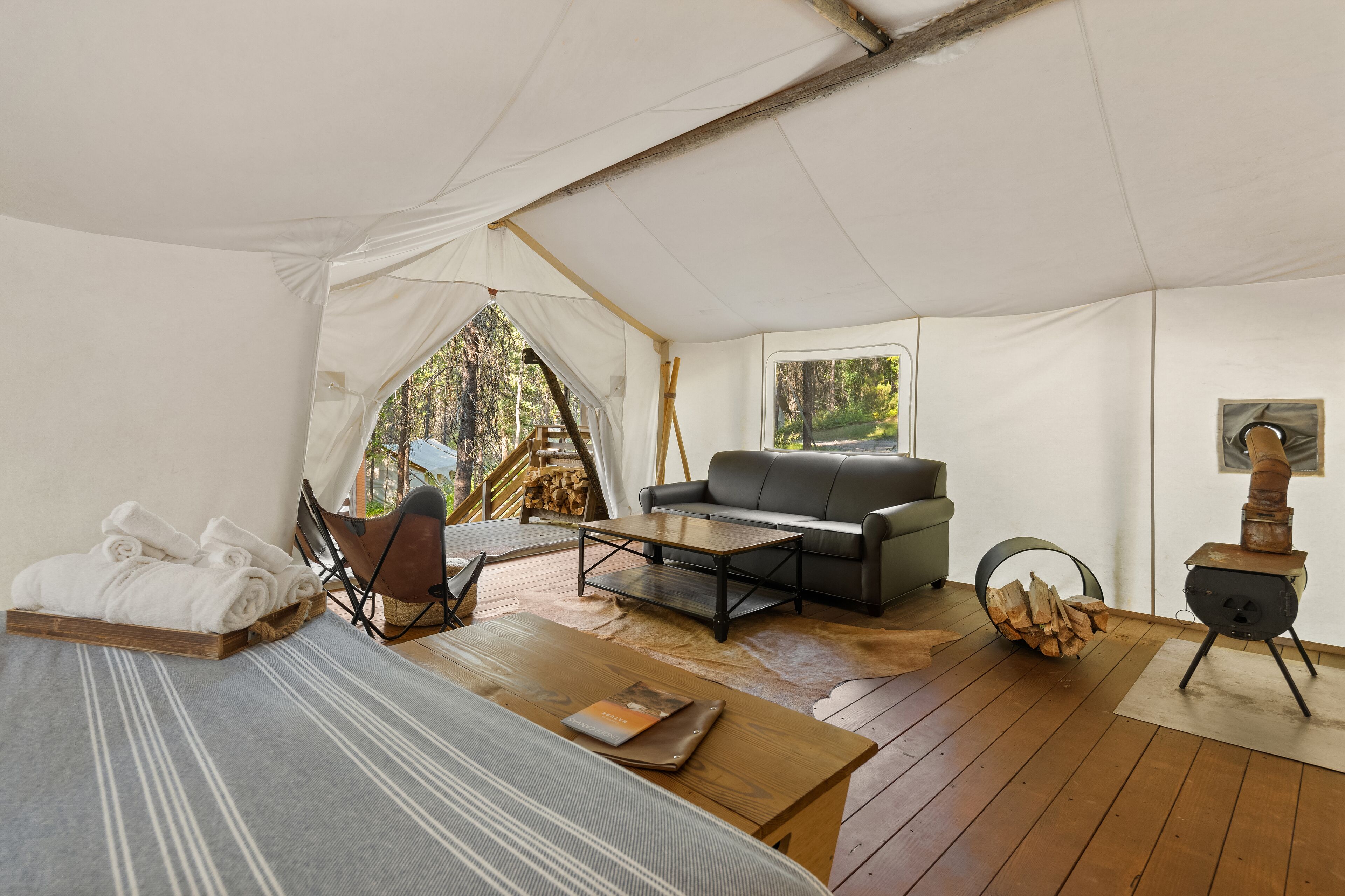 A tented accommodation with a sofa, a small table, a bed with rolled towels, and a wood stove.