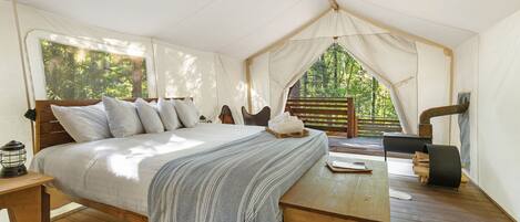 A tented room with a bed, a wooden table, and a view of the forest.