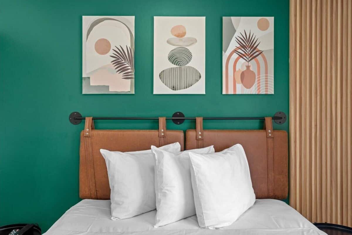 A bed with a brown headboard, white pillows, and a green wall with three abstract art pieces.