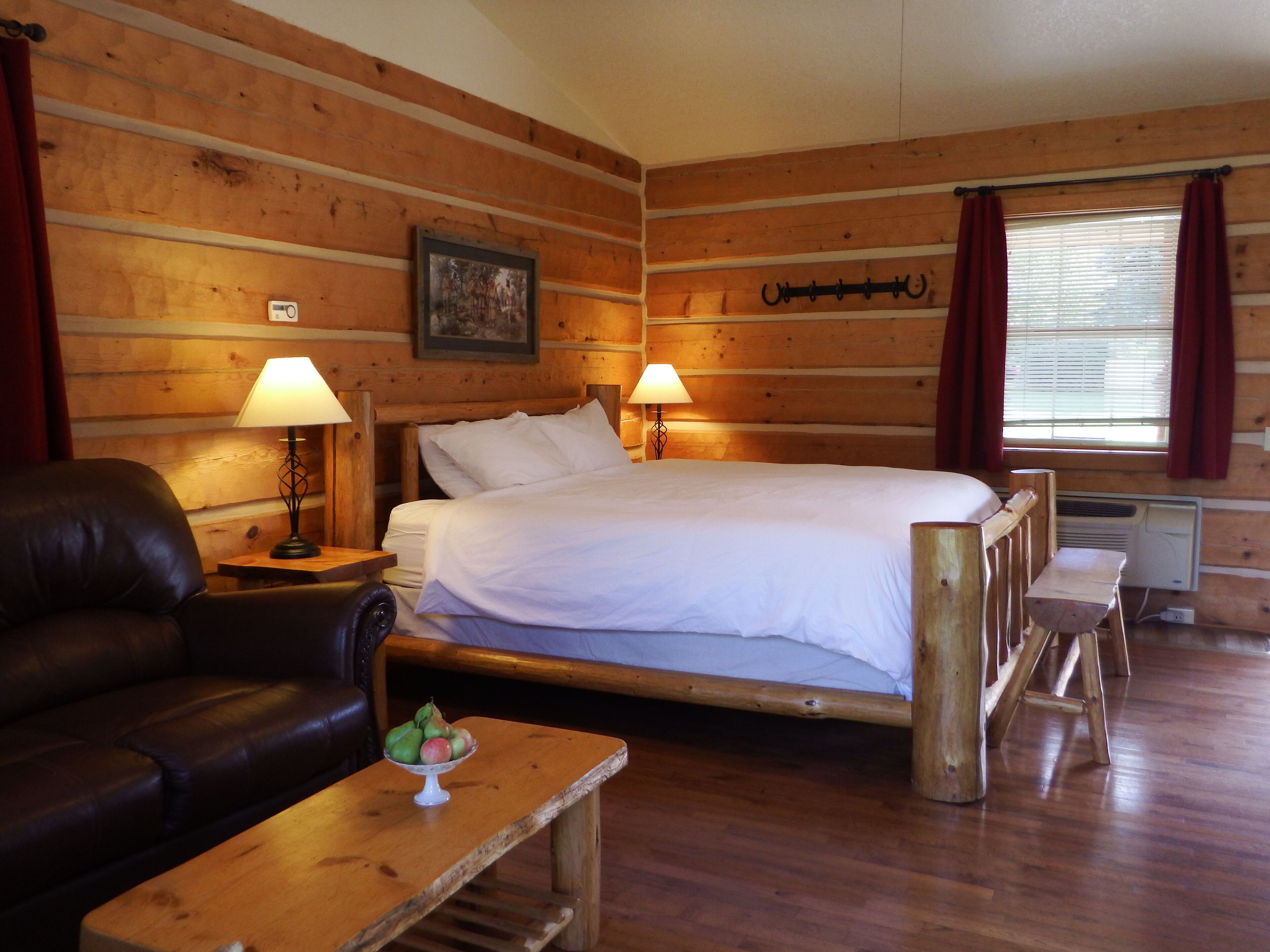 A wooden cabin room with a bed, two lamps, a leather chair, a small table with fruit, and a window with curtains.