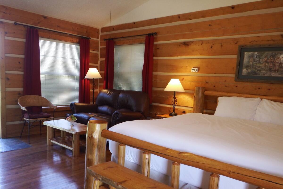 A wooden cabin room with a bed, a chair, a table, and two lamps.