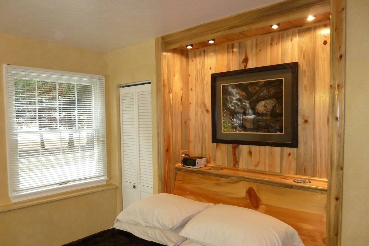 A bedroom with a bed, a window with blinds, a door, a wooden wall panel, and a framed picture.