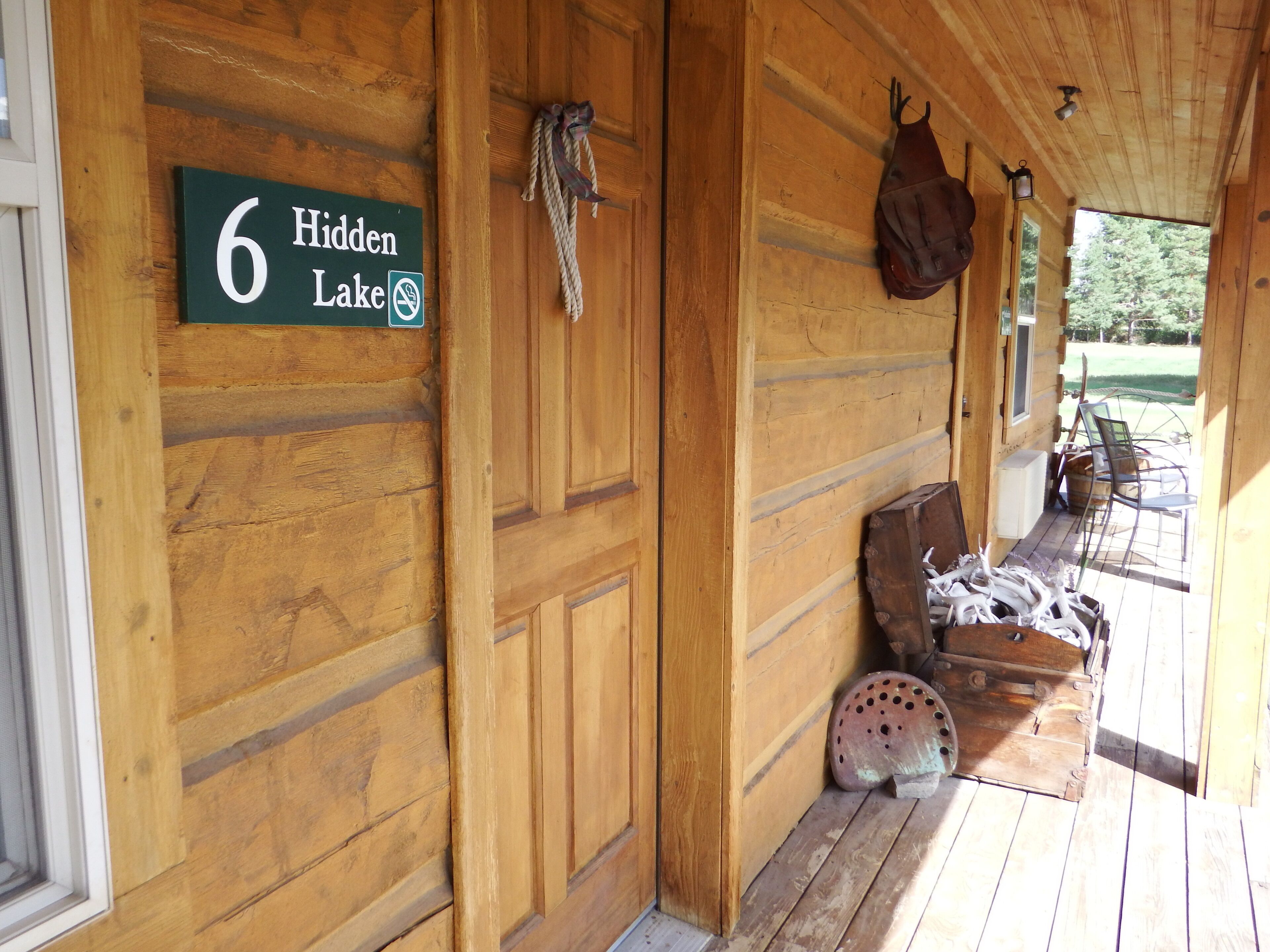 A wooden cabin with a sign reading "Hidden Lake" and a rustic porch area with outdoor seating and a large ceramic pot.