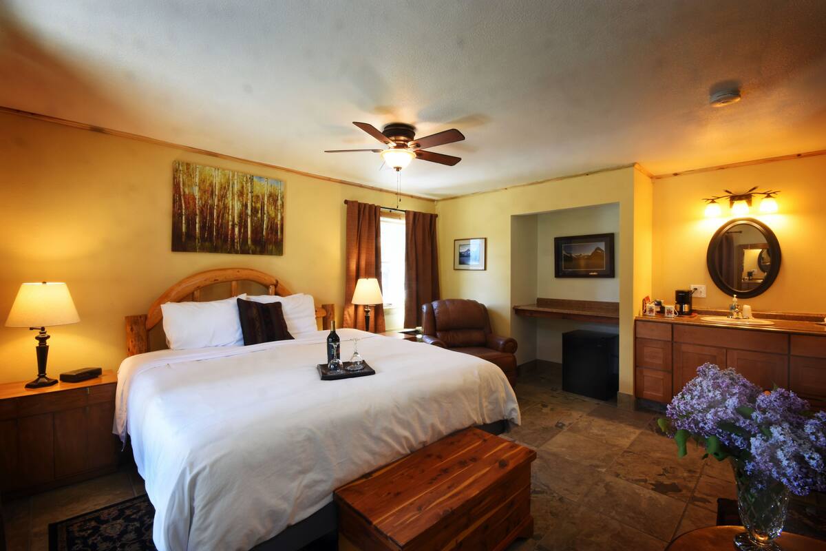 A hotel room with a bed, a wooden bench, a desk with a chair, a bathroom with a sink and mirror, and a ceiling fan.
