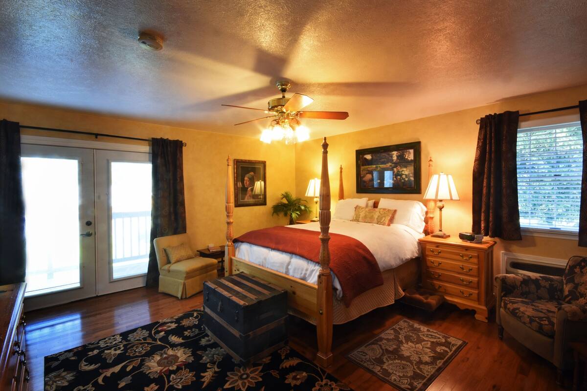 A bedroom with a four-poster bed, a ceiling fan, a nightstand with a lamp, a chair, and a window with curtains.