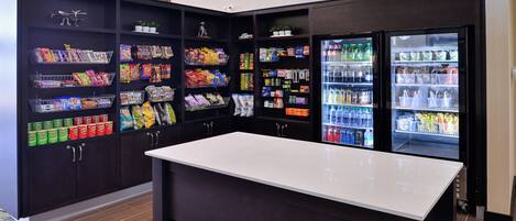 A snack bar with shelves stocked with various snacks and beverages, a table in the center, and a refrigerator on the right.