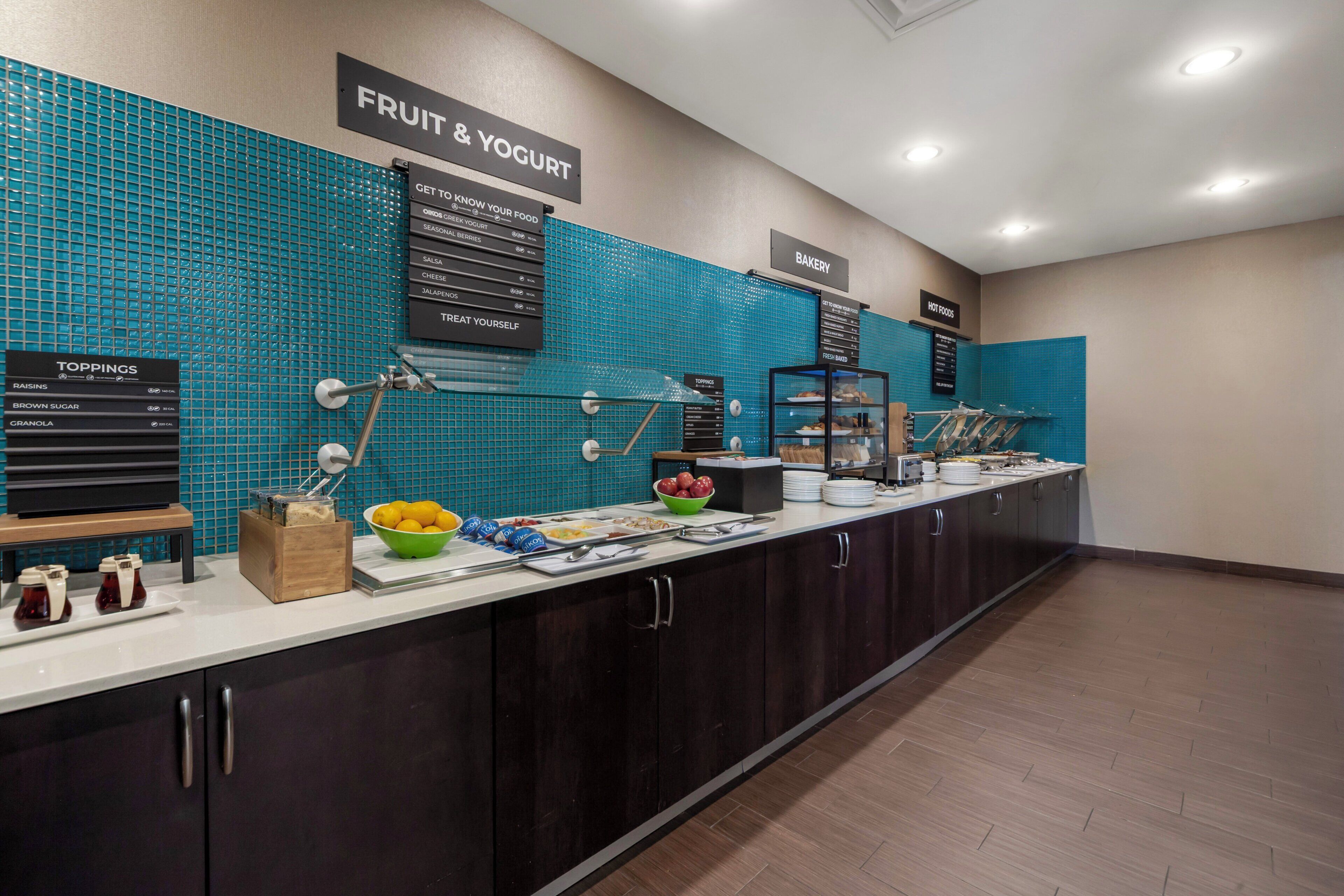 Zona de buffet con salpicadero de azulejos azules, áreas identificadas para fruta y yogur, ingredientes y alimentos calientes.