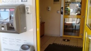 Entrada a un hotel con un mostrador de recepción automático, una puerta de cristal con un cartel y un felpudo negro.