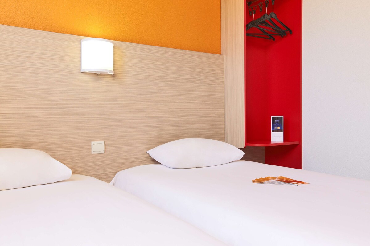 Een hotelkamer met twee bedden, een rode accentmuur, een nachtkastje met een lamp en een kapstok.