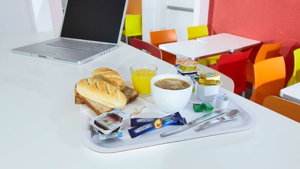Bandeja de desayuno con pan, jugo, café y condimentos sobre una mesa en una habitación de colores vivos, con una computadora portátil.